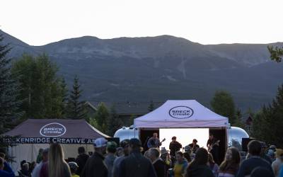 Band performing at dusk under a Breck Create tent, with an Airstream trailer, mountain backdrop, and crowd of festival-goers.