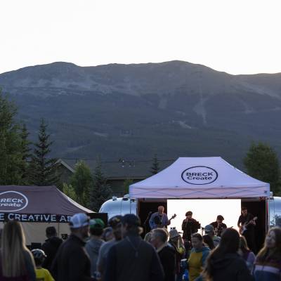 Band performing at dusk under a Breck Create tent, with an Airstream trailer, mountain backdrop, and crowd of festival-goers.