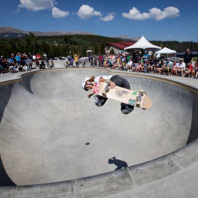 Pro skateboarder grabbing the edge of their skateboard high above the bowl while onlookers watch and a jazz band plays with the ten mile mountain range in the distance