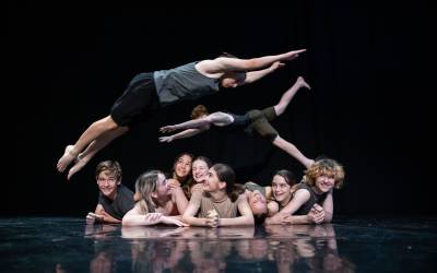 Children lay side by side on a stage floor while two other children leap over them during the Circa Youth Workshop